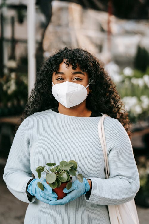 Black woman in protective mask and gloves standing with potted plant