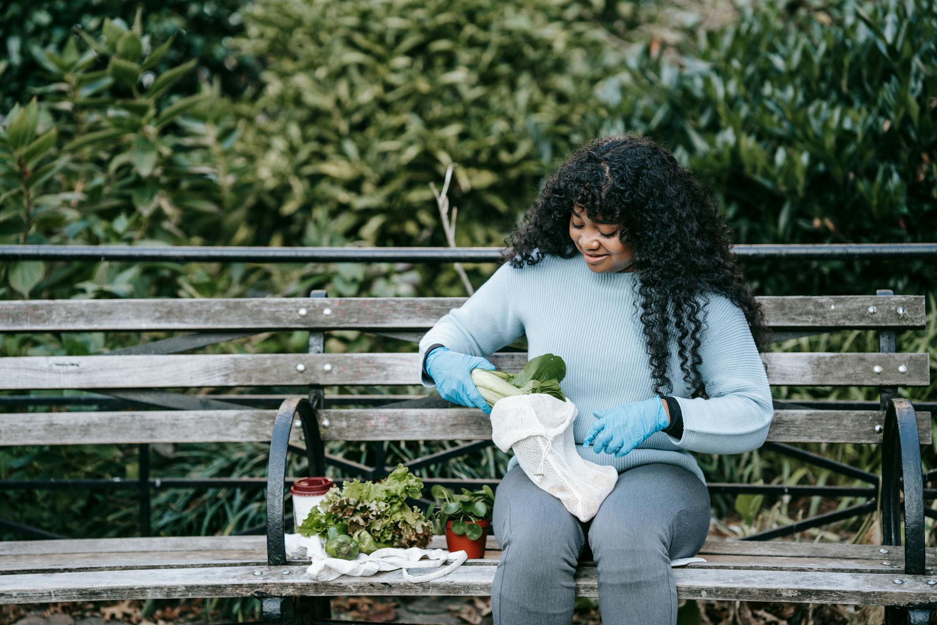 Cheerful African American woman sitting on wooden bench and putting fresh salad in eco friendly sack against green bushes in daytime