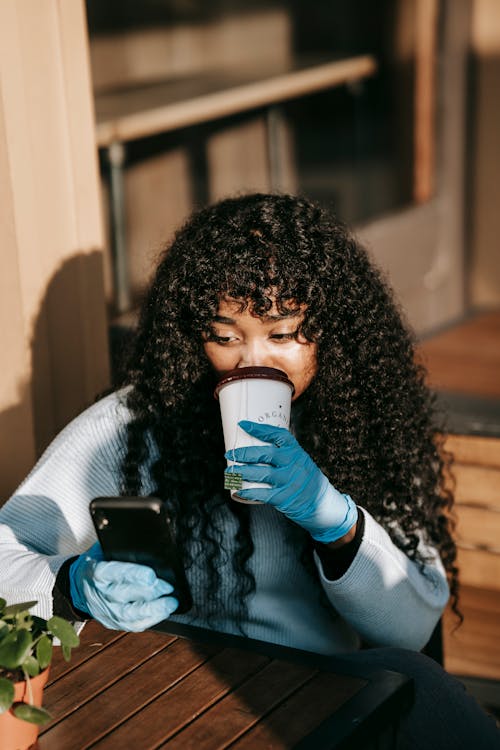 Calm African American female with long curly hair using smartphone and drinking cup of takeaway coffee while sitting at table on terrace in sunny day