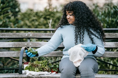 Black female sitting on bench and putting fresh vegetables in fabric sacks
