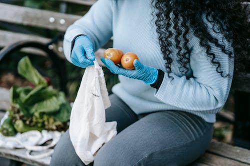 Crop anonymous female with long curly hair in casual clothes and gloves sitting on wooden bench and putting fresh vegetables in eco sack in daylight