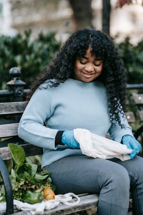 Positive African American woman with long curly hair in casual clothes sitting on wooden bench and putting vegetables in sack in garden in daytime