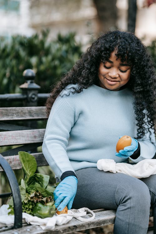 Positive young African American lady in casual clothes and gloves sitting on bench in street examining orange pumpkins near green pepper and salad leaves with reusable bags near shrubs