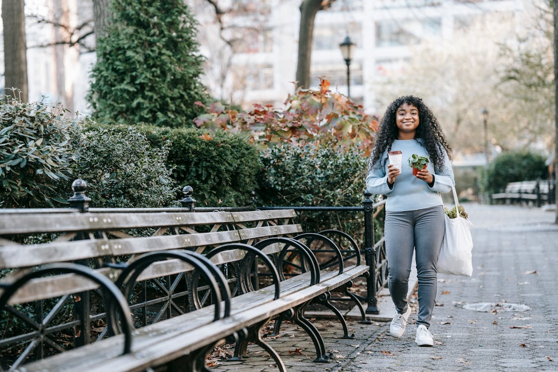 Full length of smiling young black lady strolling with shopping bag with greenery and to go cup with coffee and potted plant near bench and shrubs in daytime in park