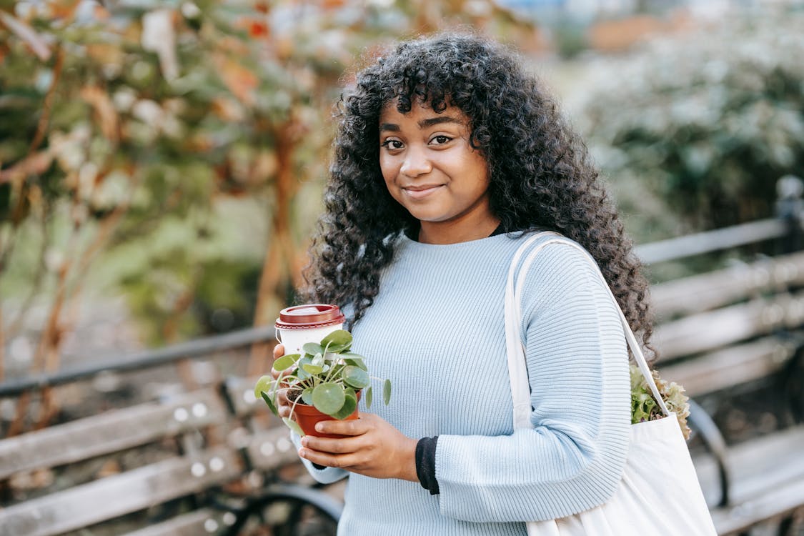 Positive young black woman standing with shopping bag with greenery and takeaway drink with potted green plant in park near bench and shrubs in daytime while looking at camera