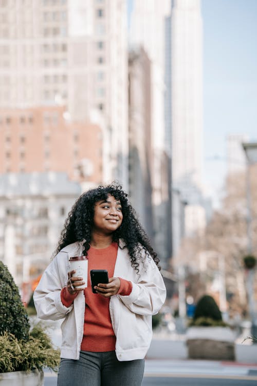 Cheerful black woman with takeaway coffee using smartphone on street ...