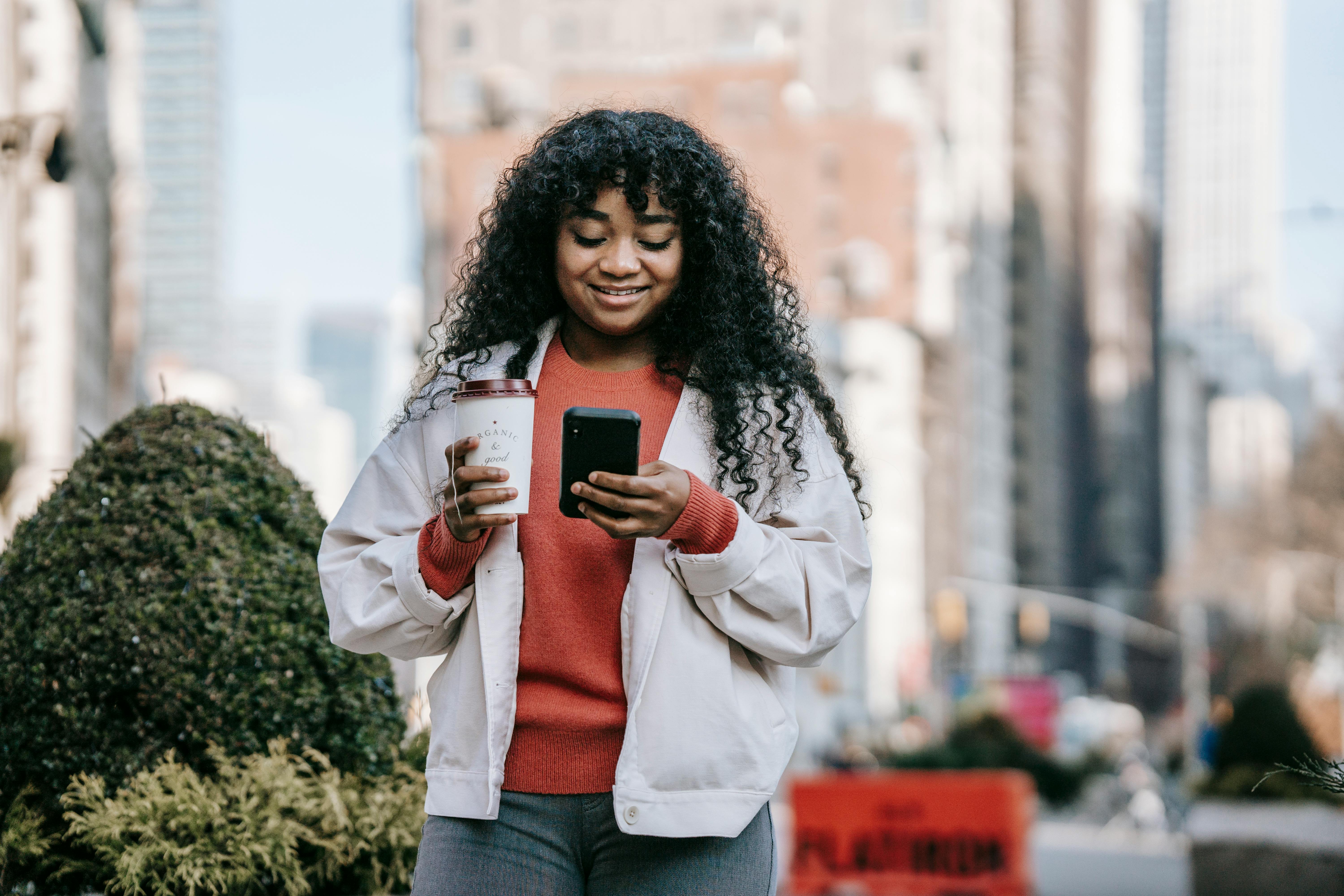 smiling black woman with takeaway coffee browsing smartphone on street