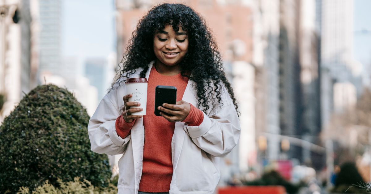A young woman smiling while looking at a budgeting app on her phone, with a coffee shop in the background