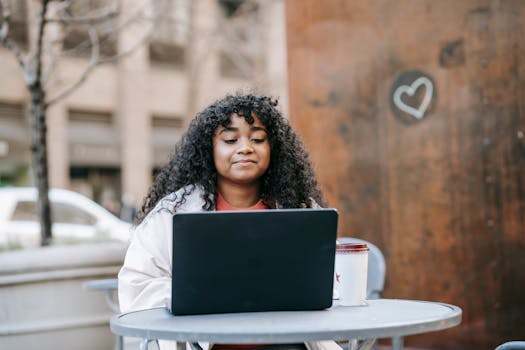 Black woman using laptop and sipping coffee at a café outdoor patio, enjoying the day.