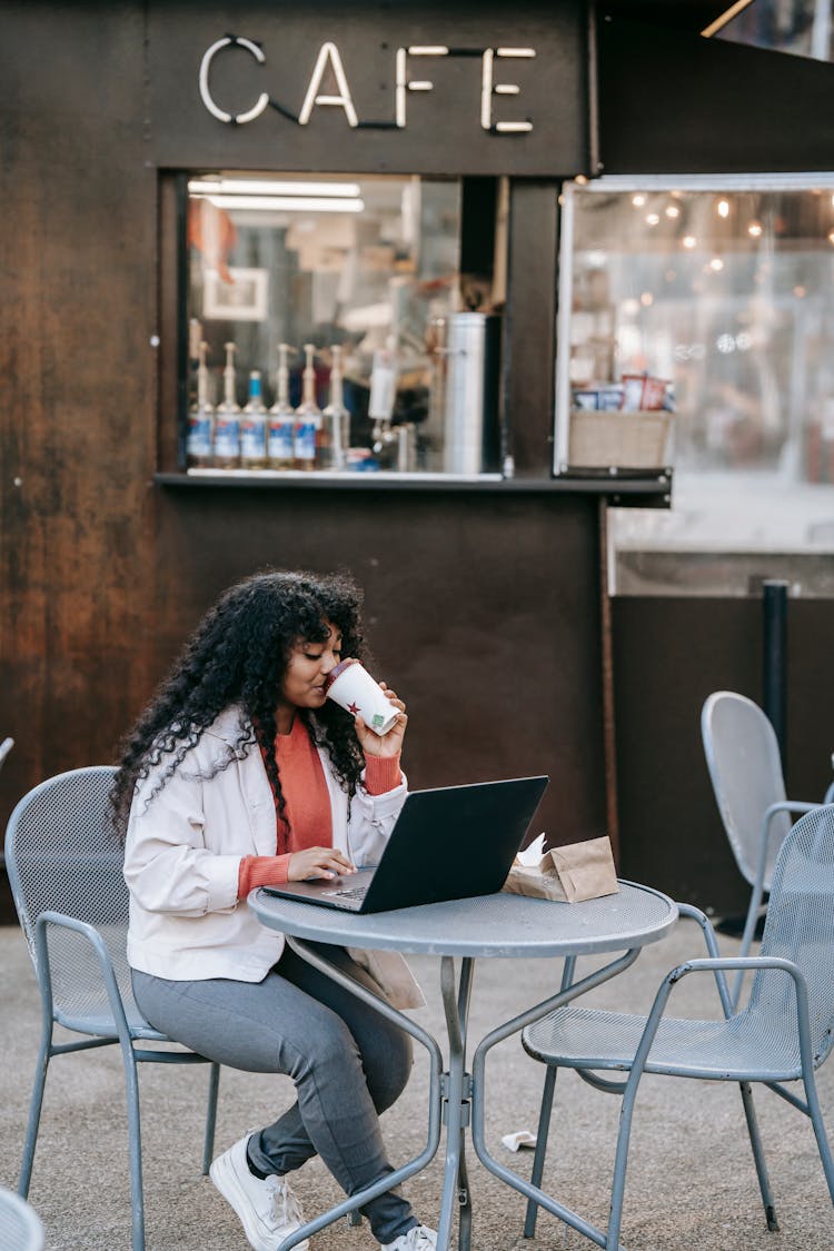 Black Woman Drinking Coffee And Using Laptop In Street Cafe