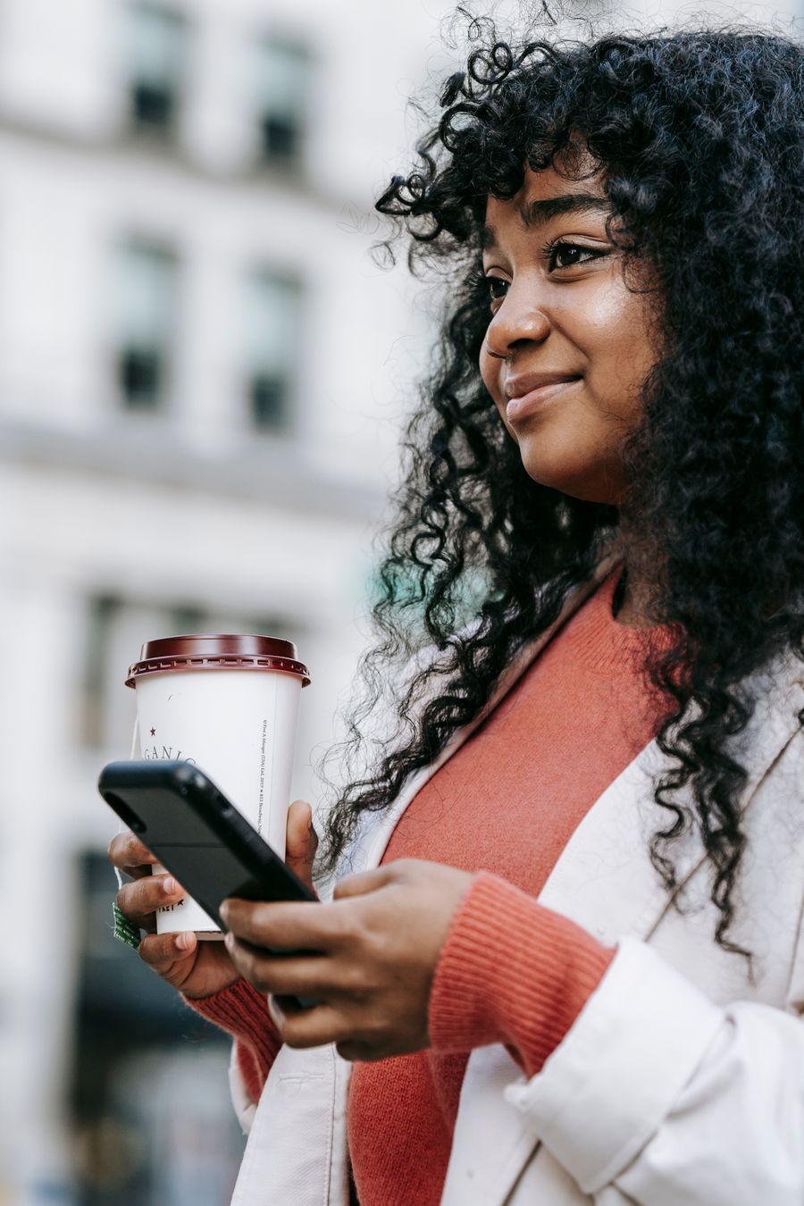 Une jeune femme consulte son smartphone en ville, un café à la main