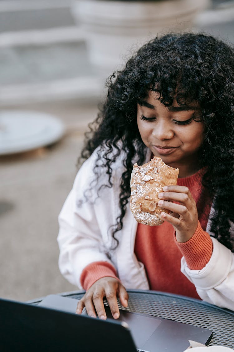 Black Woman Eating Sandwich And Browsing Laptop In Street Cafeteria