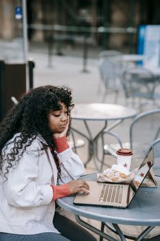 Side view focused African American female wearing casual clothes browsing netbook while sitting at table with takeaway coffee and tasty sandwich in outdoor cafeteria