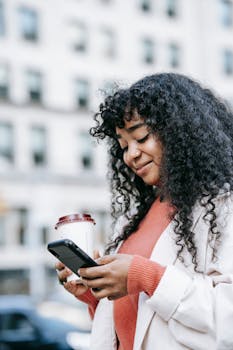 African American woman with curly hair smiles while using smartphone and holding takeaway coffee outdoors.