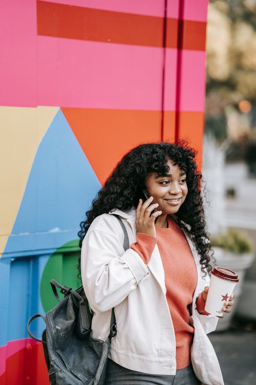 Cheerful African American female wearing casual clothes having pleasant conversation via mobile phone while standing with takeaway coffee near colorful wall on street