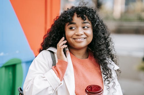 Free Happy pleasant African American female in casual outfit with takeaway coffee having conversation via mobile phone on street and looking away contentedly Stock Photo
