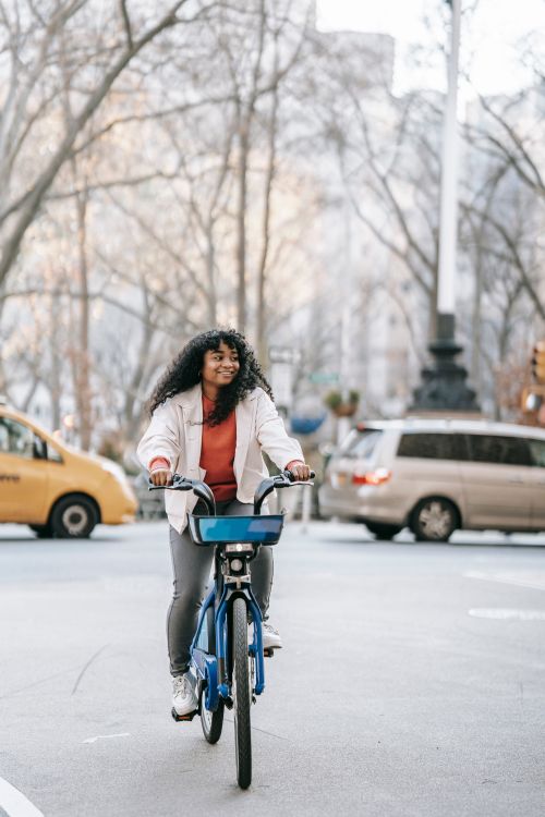 Free Full length smiling African American female in casual clothes riding bicycle on busy city street and looking away happily on early spring day Stock Photo
