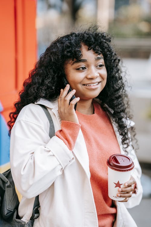 Content African American female with curly hair enjoying takeaway coffee and having conversation via mobile phone while standing on street