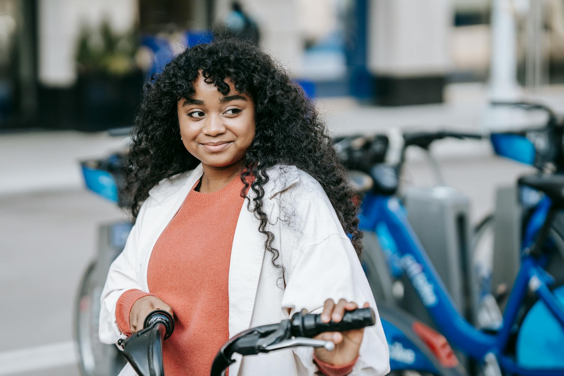 Smiling African American female cyclist wearing casual outfit standing with bicycle near bicycle sharing station on modern city street