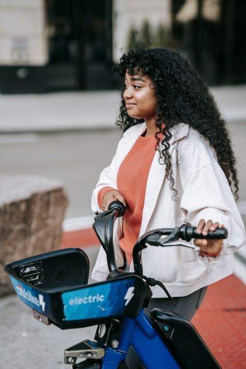 Free Positive African American female with long curly hair walking with bicycle along modern city street and looking away dreamily Stock Photo
