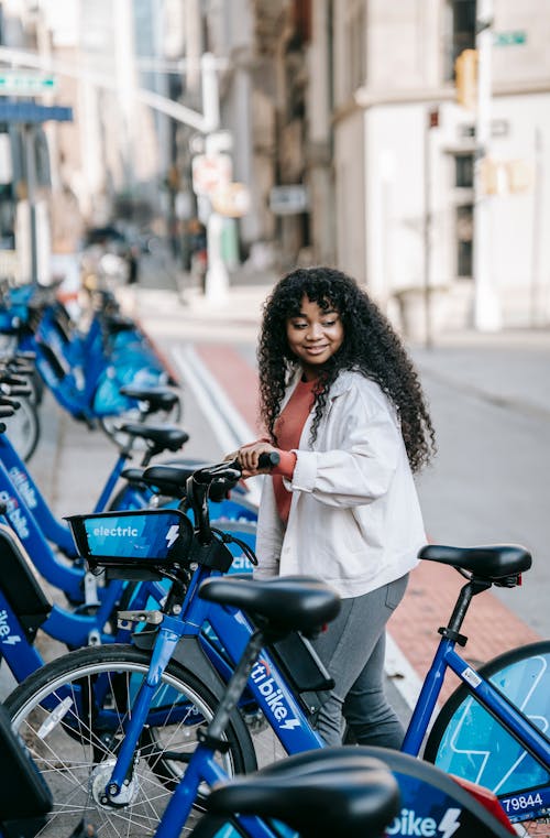 Side view glad African American female with long curly hair renting bicycle in public bicycle sharing station on modern city street