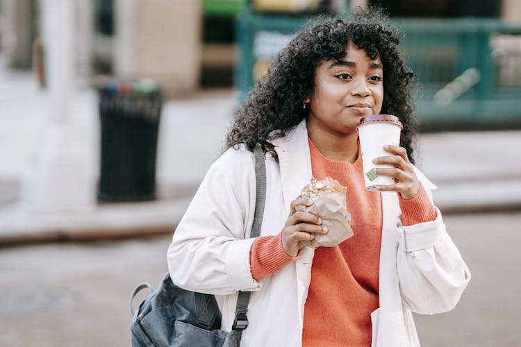 Black Woman Drinking Takeaway Coffee And Eating Sandwich On Street