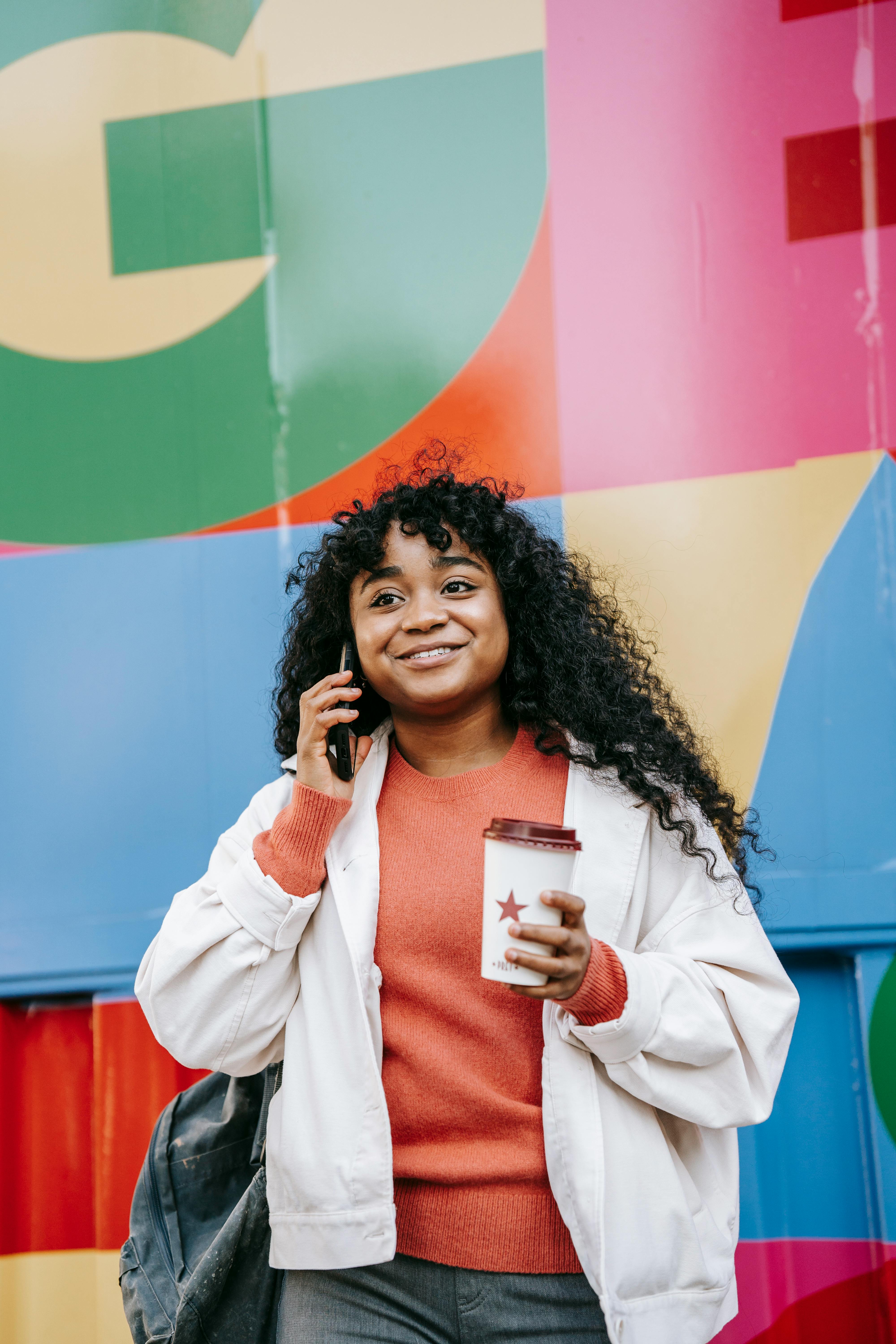 smiling black woman with takeaway coffee talking on smartphone