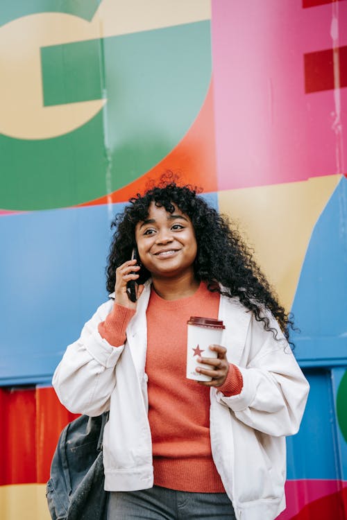 Cheerful African American female in casual wear with takeaway coffee having conversation via mobile phone while standing against colorful wall on street