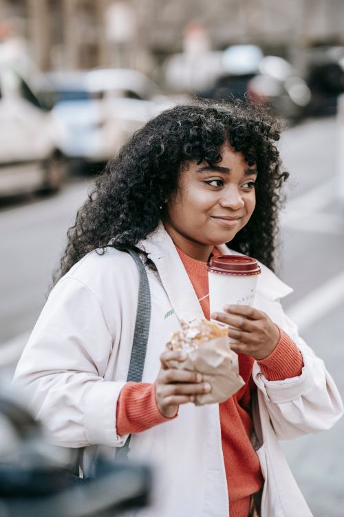 Free Cheerful African American female in casual outfit enjoying hot takeaway coffee and tasty sandwich while walking on busy city street Stock Photo