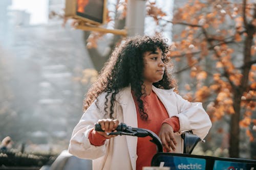 Free Dreamy young black lady in casual outfit looking away on street with bike in town in sunny day near tree with orange leaves and traffic light Stock Photo