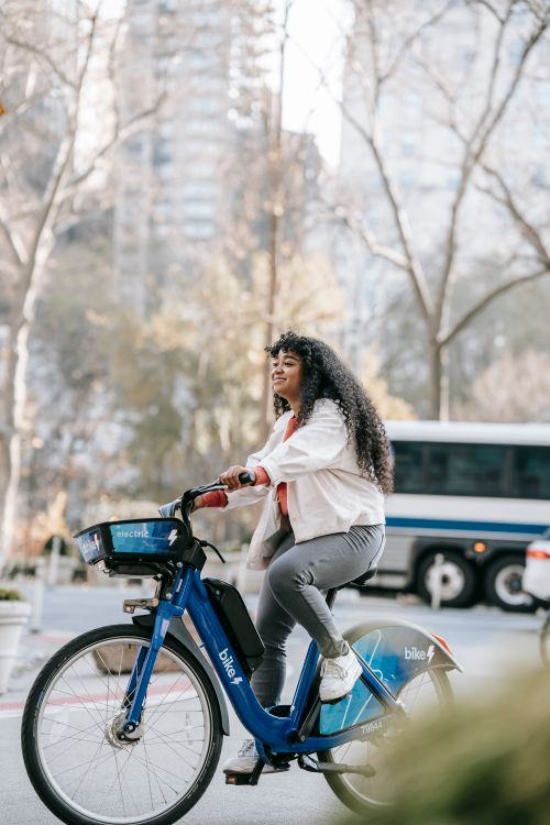 Free Full body African American lady wearing casual clothes while riding bike on street and looking away in town in sunny day near road with bus and buildings near trees Stock Photo
