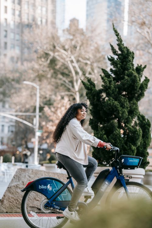 Free Side view full length of young black lady in casual outfit riding bicycle on road in town on street near trees and buildings in sunny day Stock Photo