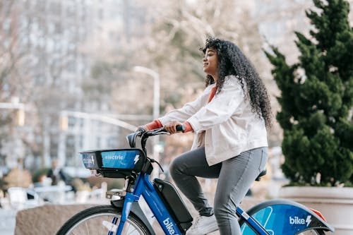 Free Side view of positive African American lady wearing casual clothes while riding bike in town in park in sunny day near buildings and trees Stock Photo