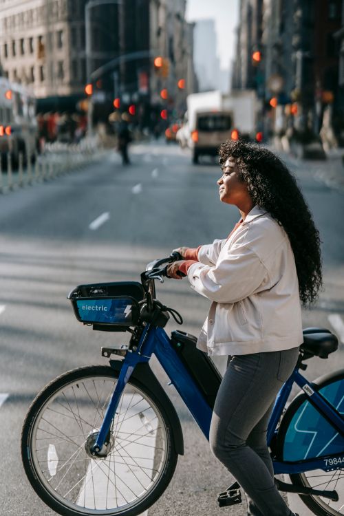Free Side view of young African American lady wearing casual clothes while walking on crosswalk with bike in city street in sunny day near buildings and vehicles on road Stock Photo