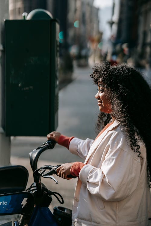 Side view of young black lady in casual outfit standing on road in street near pole with bicycle in town in sunny day near buildings