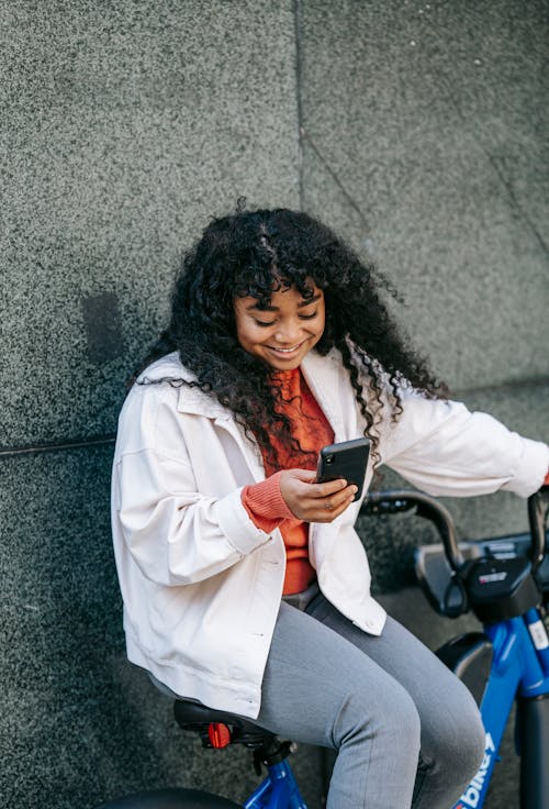 Positive black woman sitting on bicycle with phone near fence