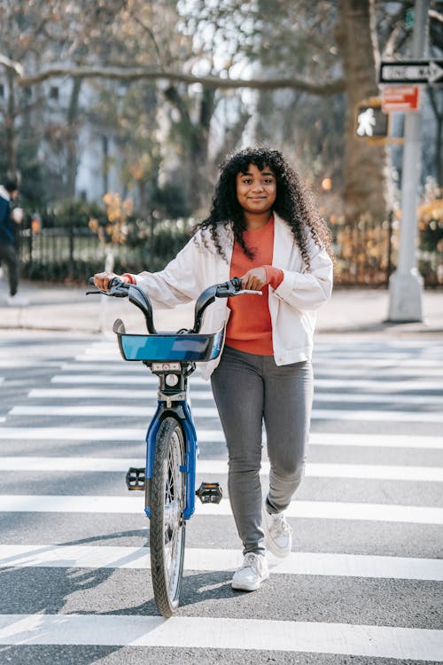 Free Full body of smiling young black lady in casual outfit walking on crosswalk with bicycle near road in street in town in sunny day near buildings while looking at camera Stock Photo