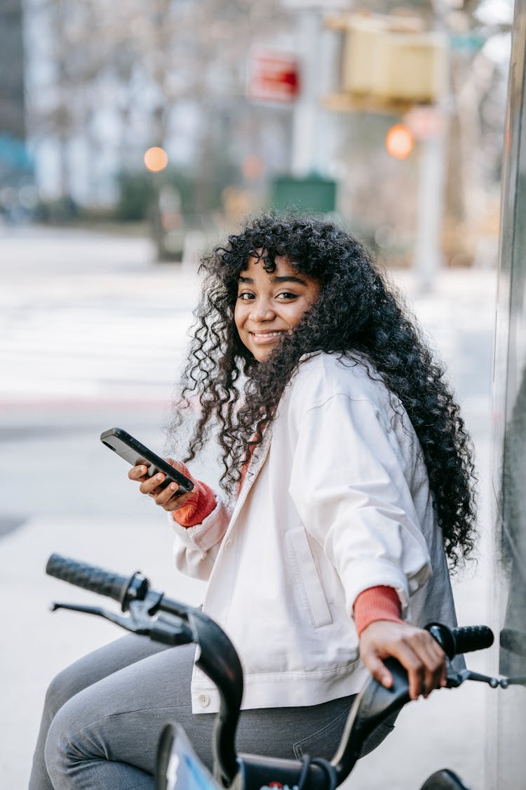 African American Female With Smartphone Sitting On Bike In Street