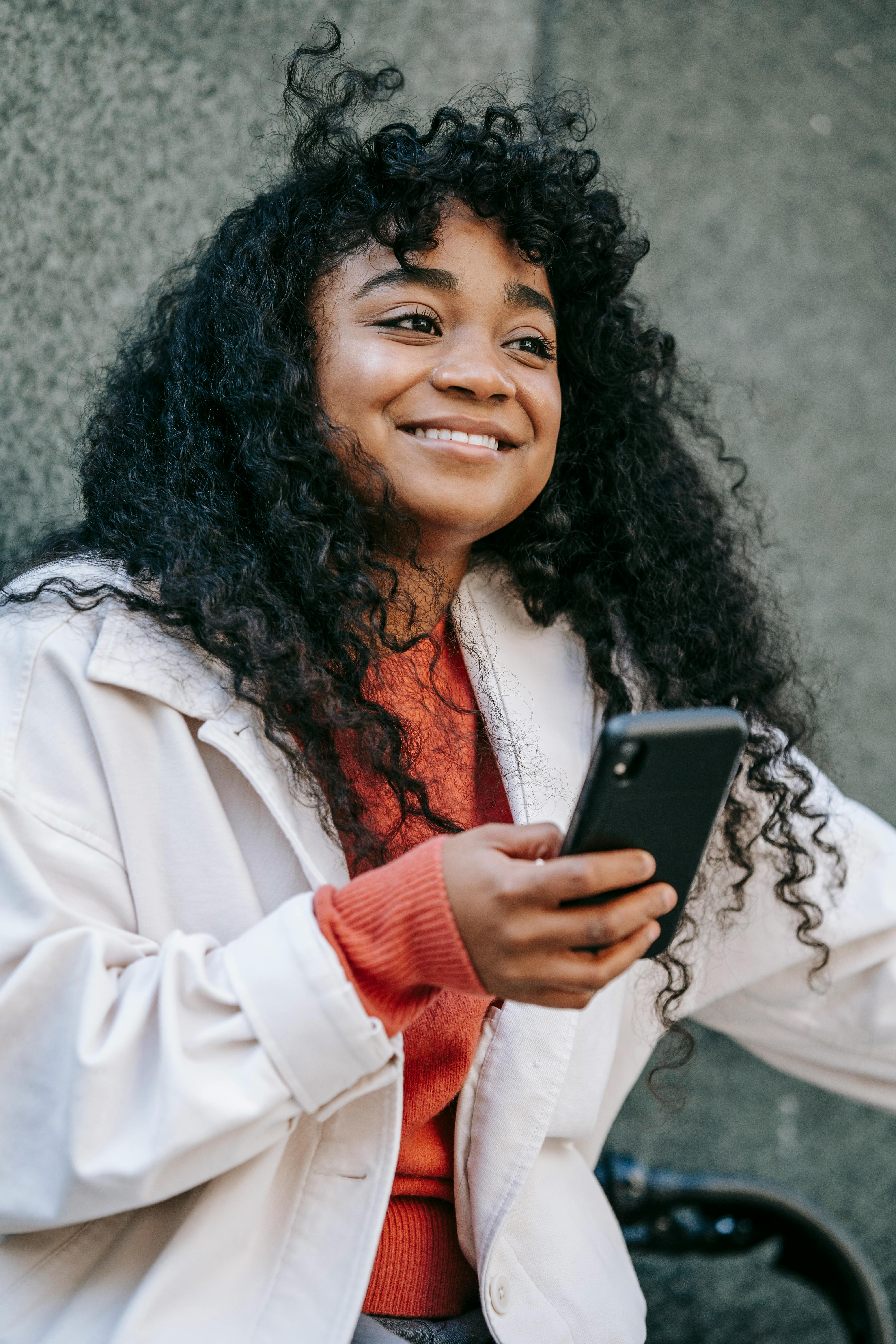 happy black lady sitting on bicycle with cellphone near fence