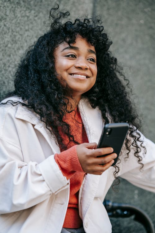 Positive young African American female wearing casual clothes near bike and texting on phone in daytime in city street near building wall and looking away