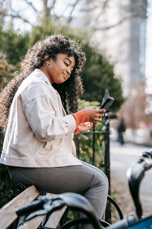 Side view of young African American female wearing casual clothes while sitting on bench and text messaging on cellphone near bike and green bushes behind fence in city street