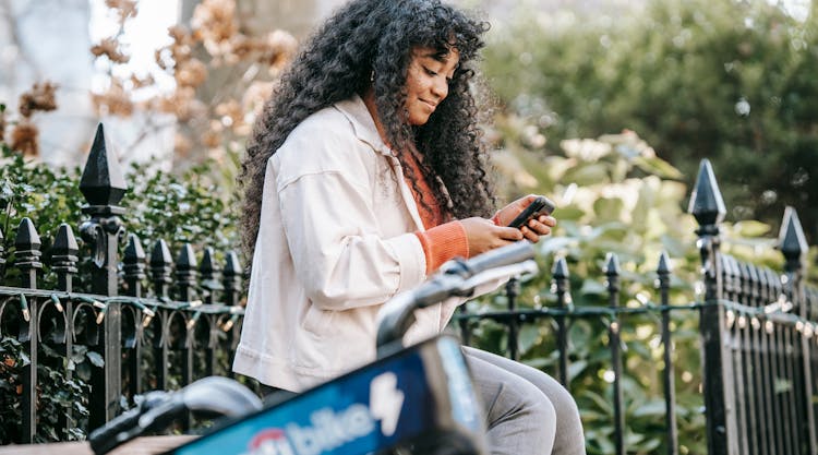 African American Female With Smartphone Sitting On Bench Near Bike