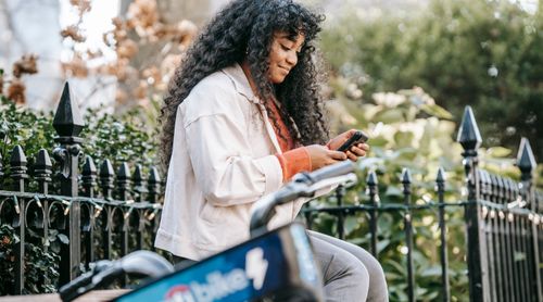 Free Side view of positive young black lady in casual outfit sitting on bench near bicycle in park while surfing mobile near fence with plants in town in sunny day Stock Photo