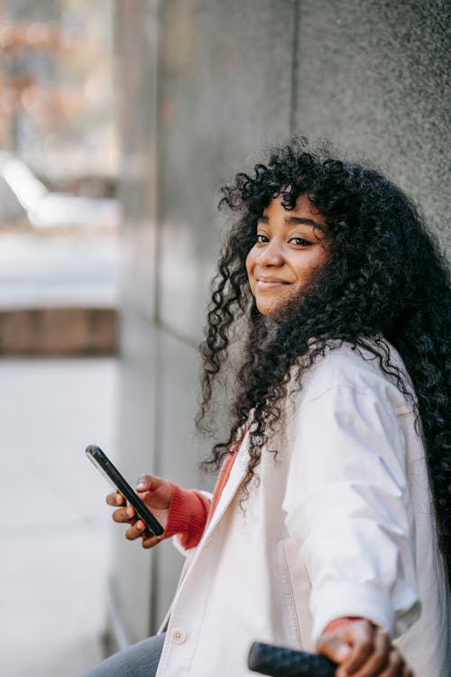 Positive young African American female wearing casual clothes while sitting on bike and texting on cellphone near building wall in city street in daytime and looking away