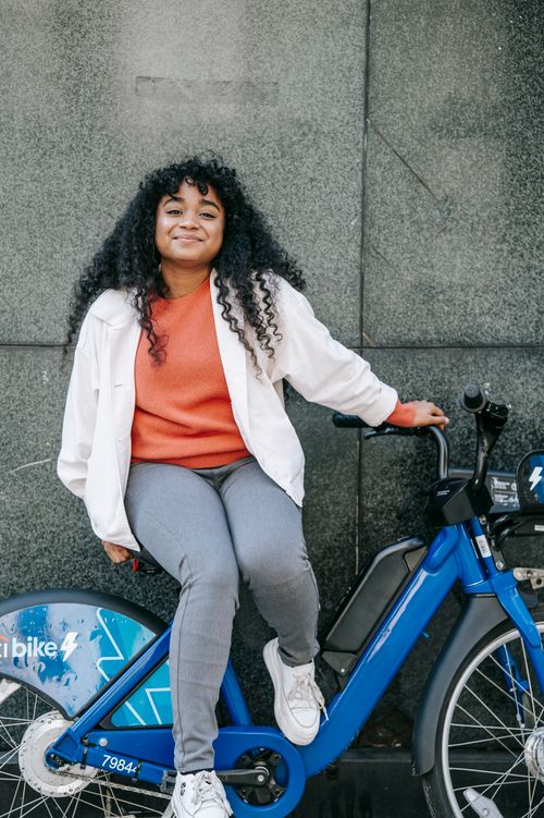 Free Cheerful African American female with long curly hair sitting on bicycle against building wall and looking at camera happily Stock Photo