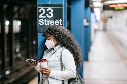 Focused African American female wearing casual outfit and protective mask standing on New York underground platform with takeaway coffee and using mobile phone