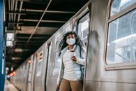 Young African American female passenger in casual clothes and mask drinking takeaway coffee and getting off train in subway station