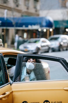 Young woman wearing a mask exits a yellow taxi on a bustling city street, reflecting cautious travel in the new normal.