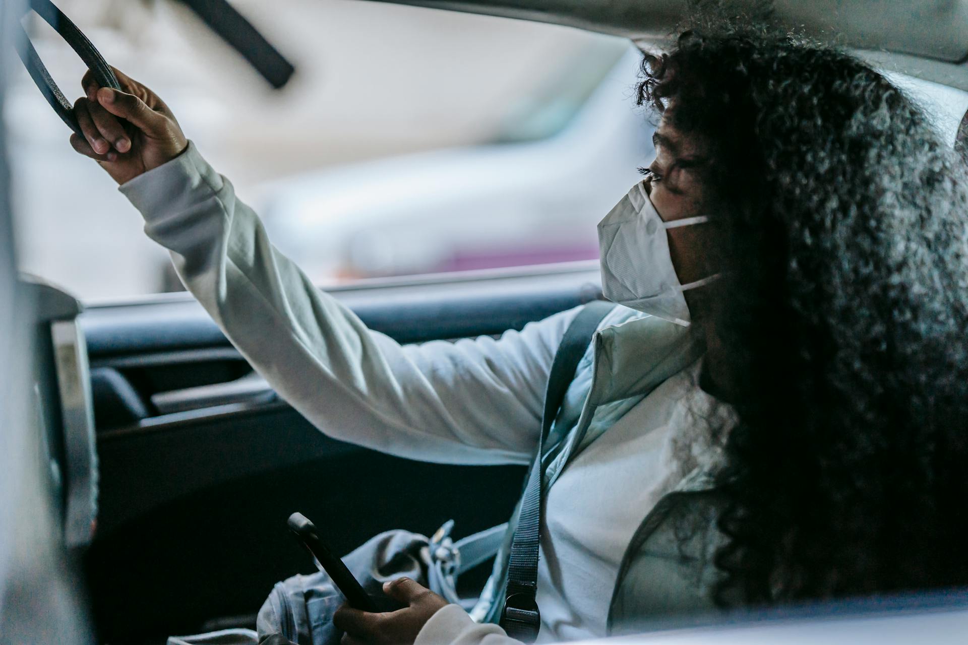 Side view of anonymous young African American female passenger in casual clothes and protective mask holding handle and looking away while sitting on backseat of taxi with smartphone in hand