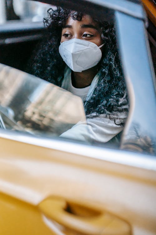 Calm black female in mask sitting in yellow taxi and looking away